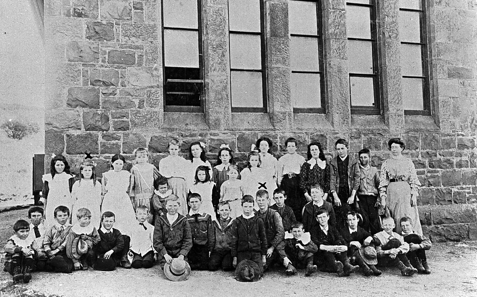 About forty-five children in three rows against the wall of a big stone building with tall sash windows: girls in white pinafores and hair bows at the back, boys in sailor collars kneeling and sitting in front with their hats, and their teacher, a woman in a high-collared blouse, at the right.