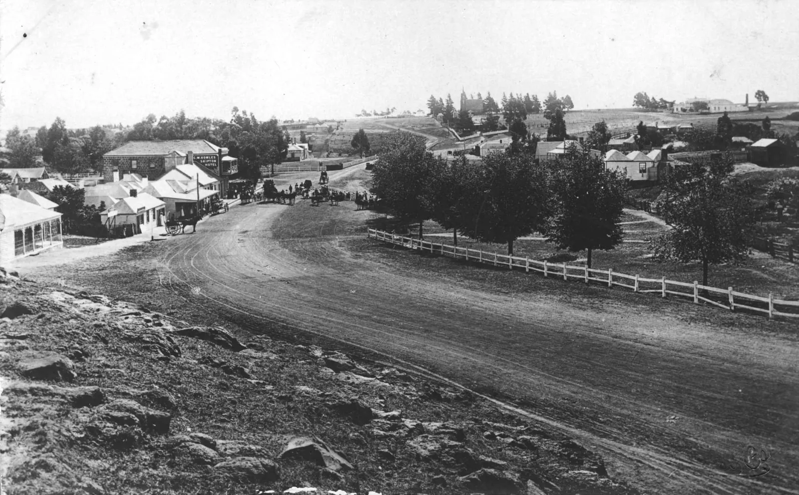 A wide dirt road sweeping down in a curve into a country town: single-storey shops with verandahs and a two-storey hotel with a big sign on the left, a crowd with buggies and saddle horses further down, a white post-and-rail fence round young trees on the right, and cottages and a church spire among pines on the hill beyond.