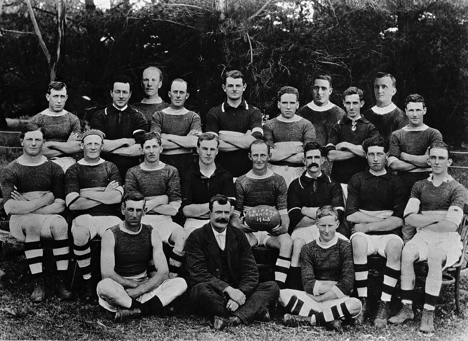 A football team in three rows against dark pines: men standing with arms folded in hooped guernseys, more seated in shorts and hooped socks, and in front a player cross-legged, a moustached man in a dark suit and a fair-haired boy, with a ball chalked Skipton Premiers 1923.