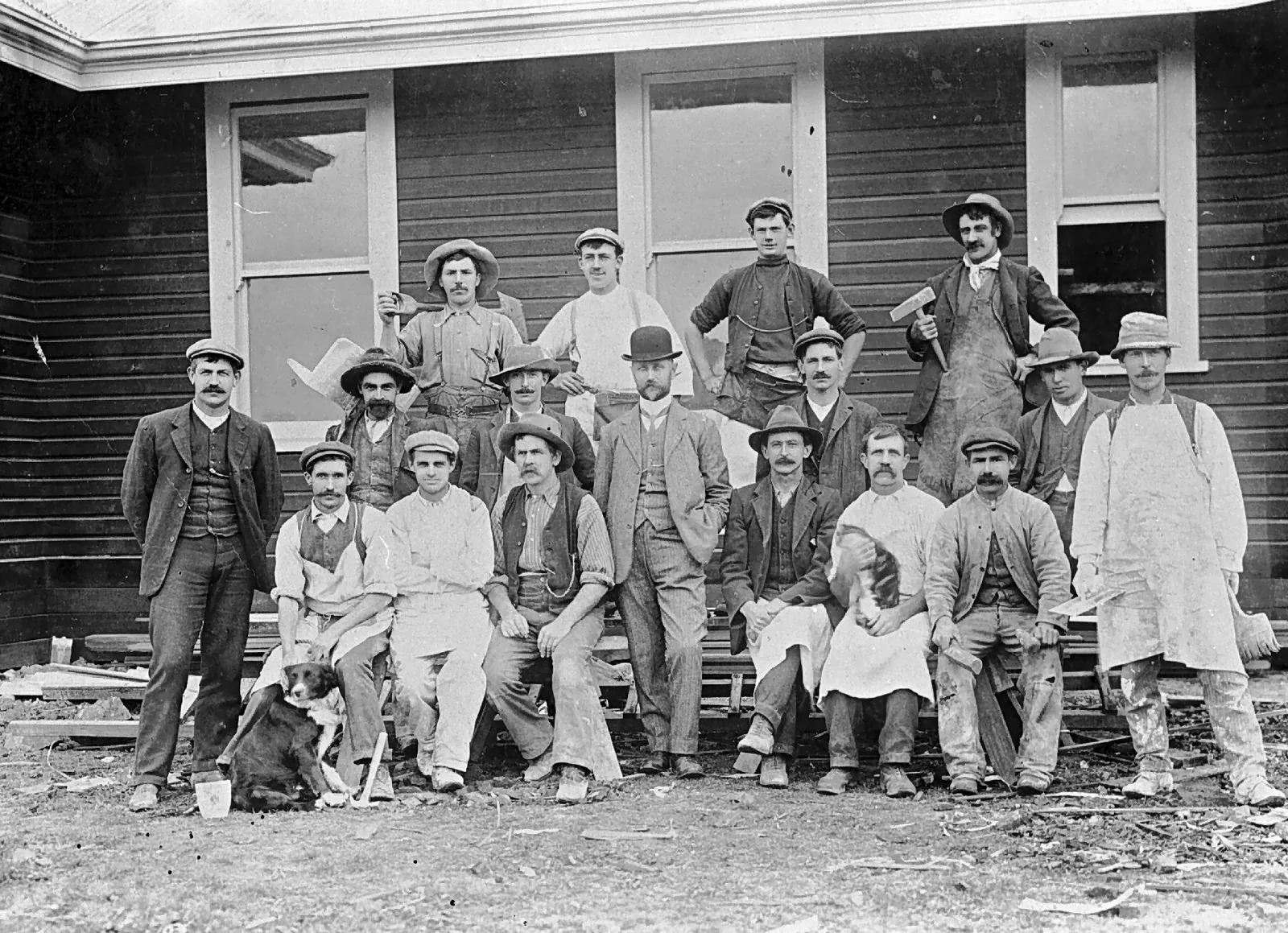 Seventeen men in three rows in front of a new weatherboard building, offcuts on the ground: tradesmen in work shirts, waistcoats and aprons holding hammers and a trowel, a man in a bowler hat and pale suit in the middle, a plasterer in a spattered white coat, a man holding a tabby cat, and a black-and-white collie at the front.